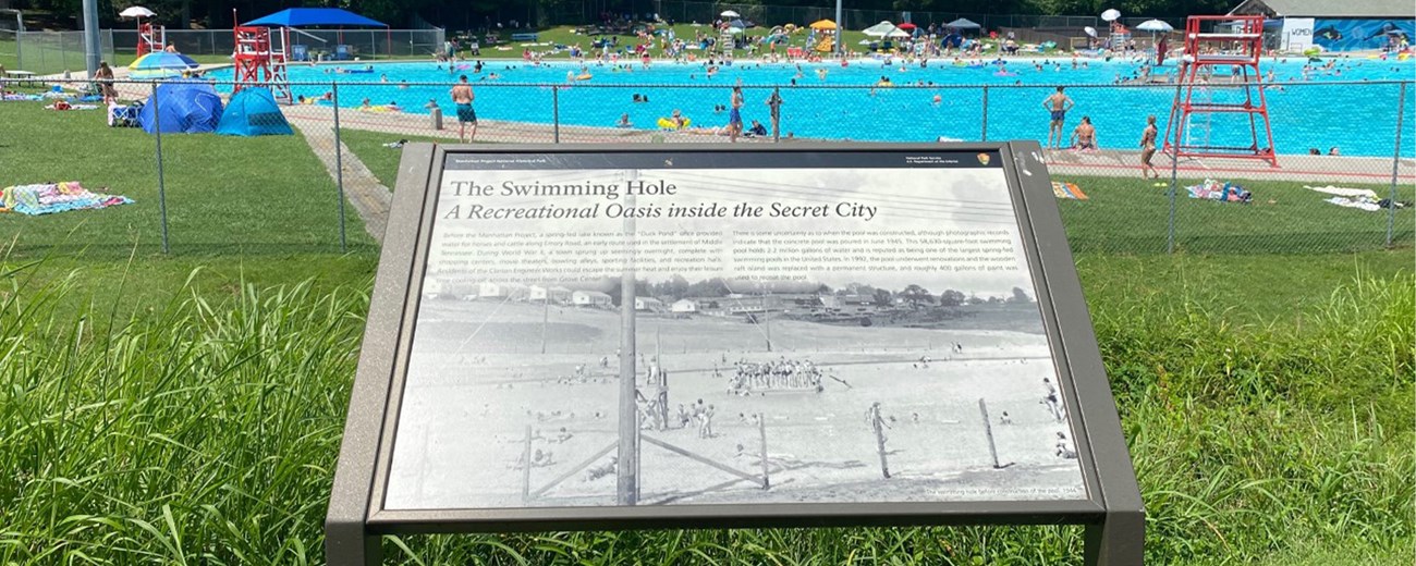 A wayside exhibit in the foreground with a busy public swimming pool behind it.