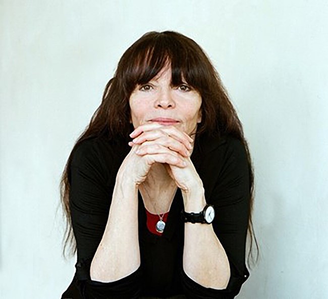 A women with long dark hair is seated resting elbows on knees and hands interlaced in front of chin looks directly at the camera