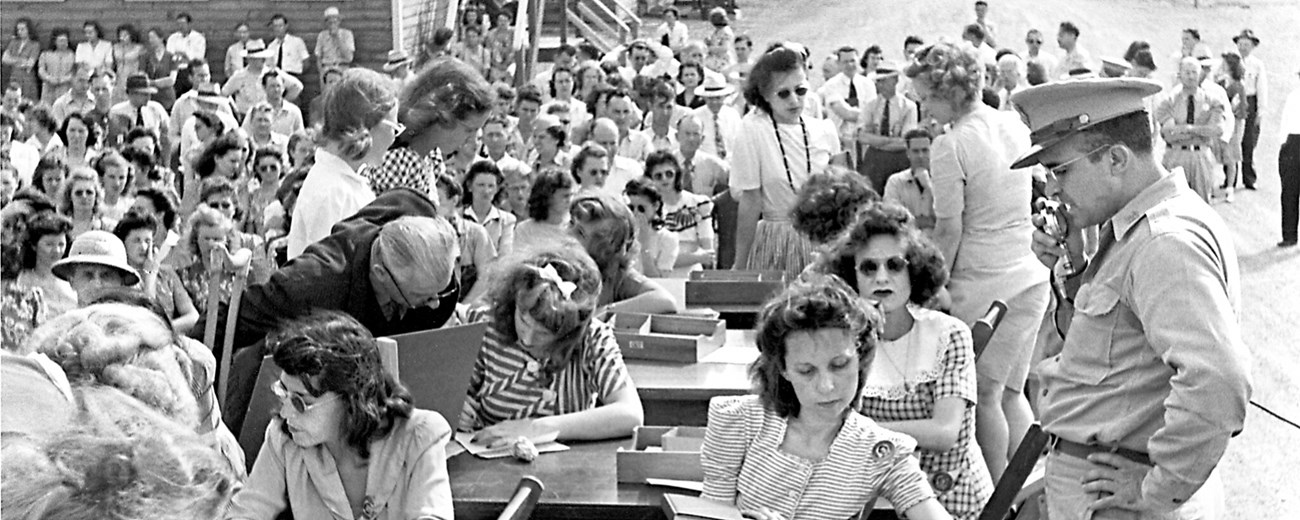 Several men and women seated at outdoor desks with military personnel looking on.