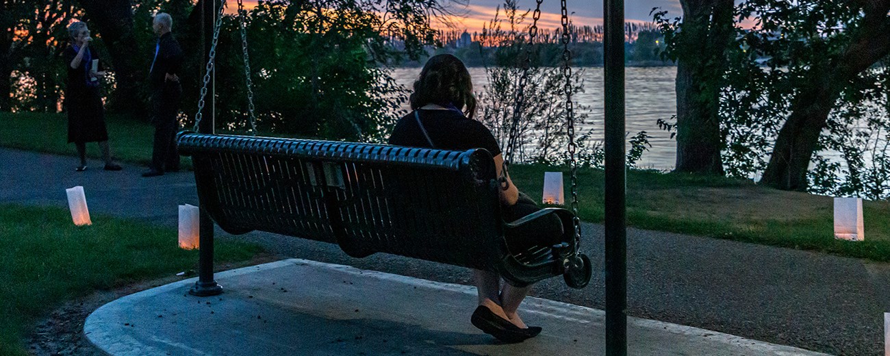 A woman sits on a swinging bench in front of the river. Luminarias line the path.