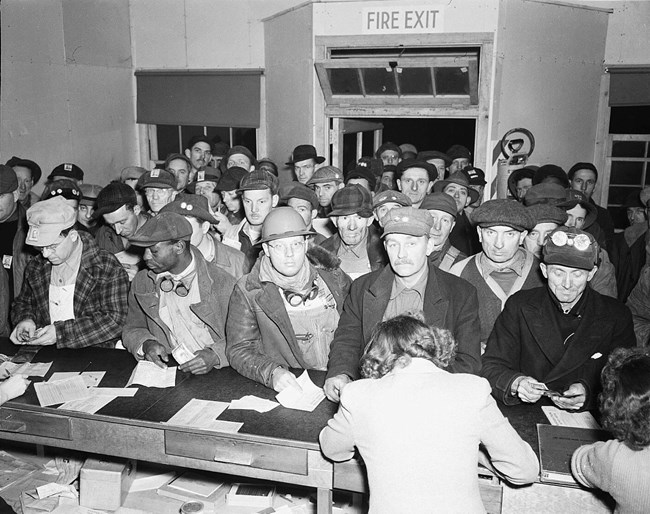 A black and white photo of a large group of men in hats waiting at a counter. There are women behind the counter.