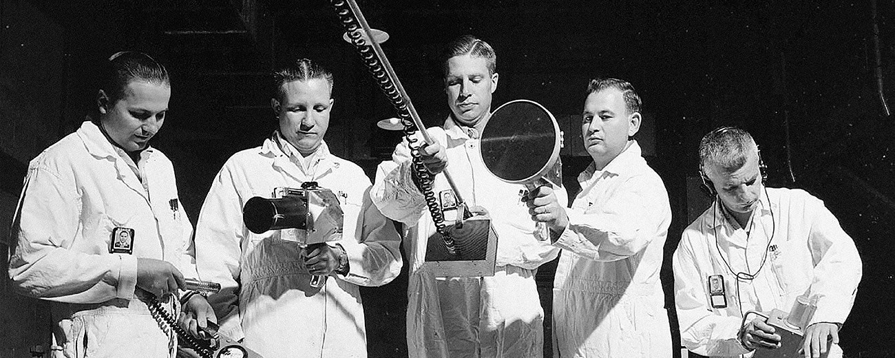 Black and white photo of five people in white coveralls holding a variety of handheld radiation detectors.