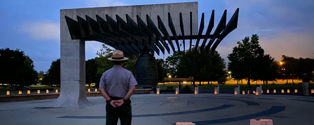 Park ranger faces large bronze bell overhanging a courtyard encircled by glowing luminaria bags.