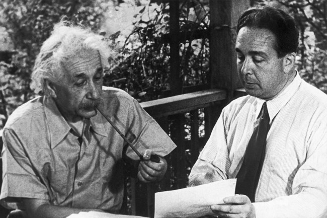A man with a long pipe on the left is sitting next to a man in a shirt and tie. They are both looking at a piece of paper on a porch with foliage in the background.