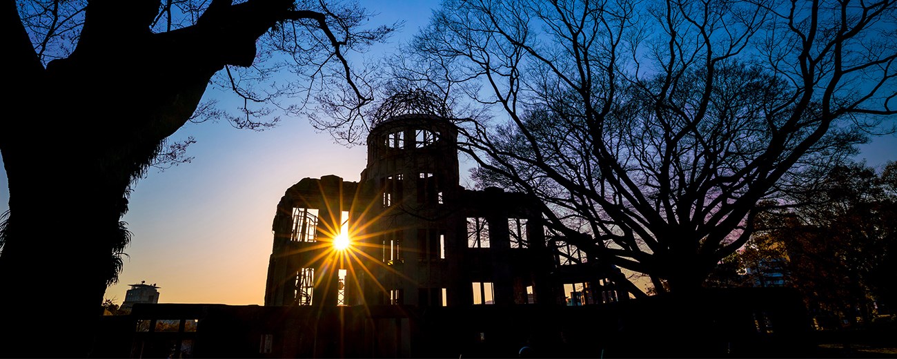 The silhouette of a domed building in ruins with the sun shining through a missing window. Trees are on either side.