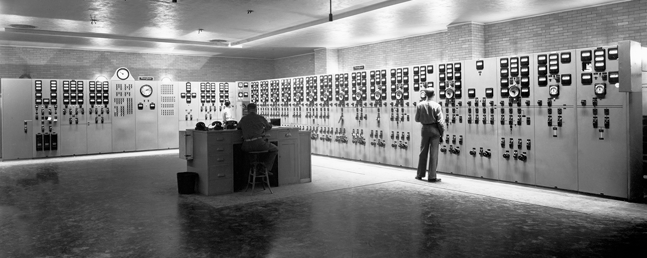 Black and white photo of a large room. A man sits at a counter in the center while another looks at dials on the wall.
