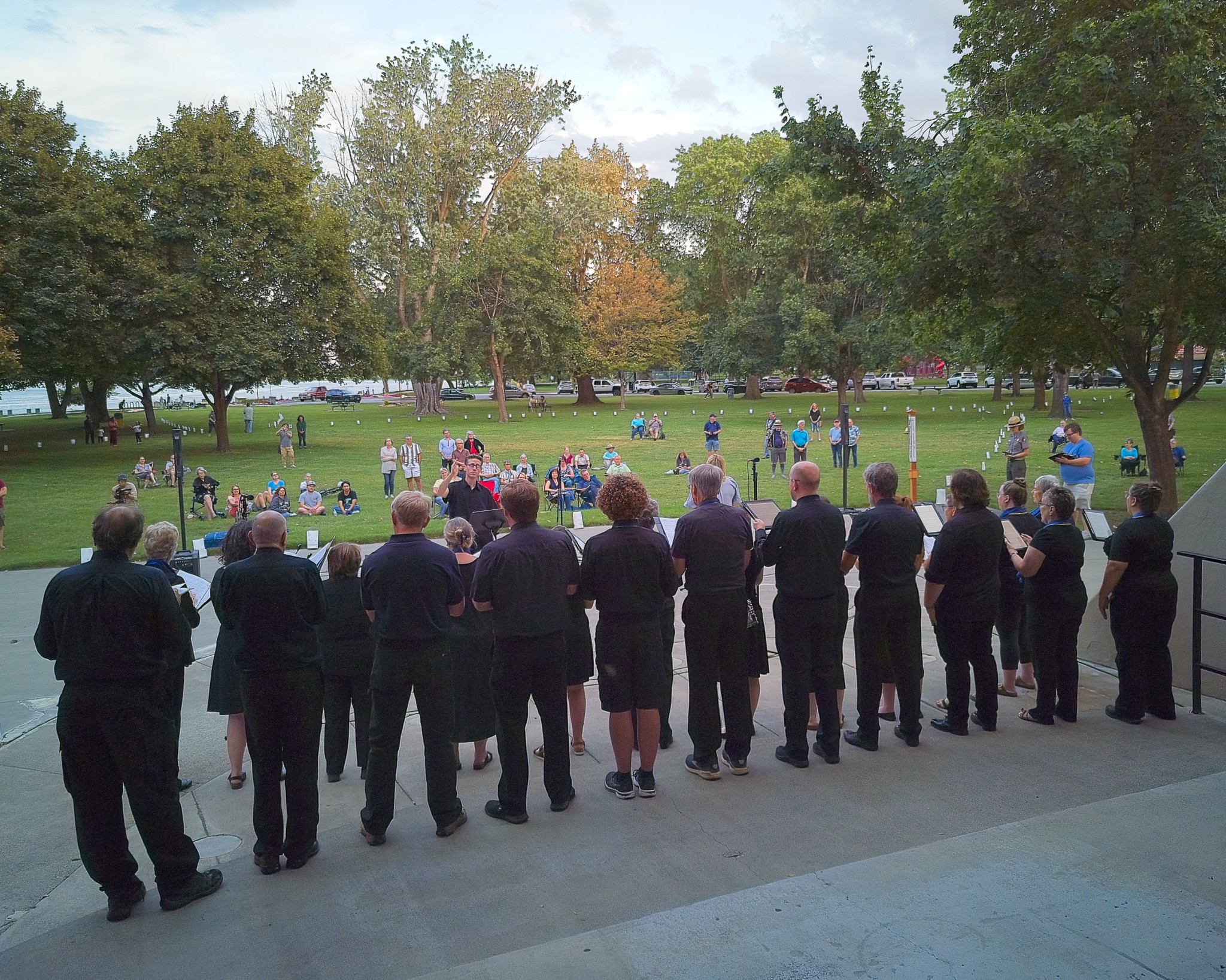 16 adults wearing black outfits stand facing a crowd of about 50 people seated and standing in a park lined with small white bags.