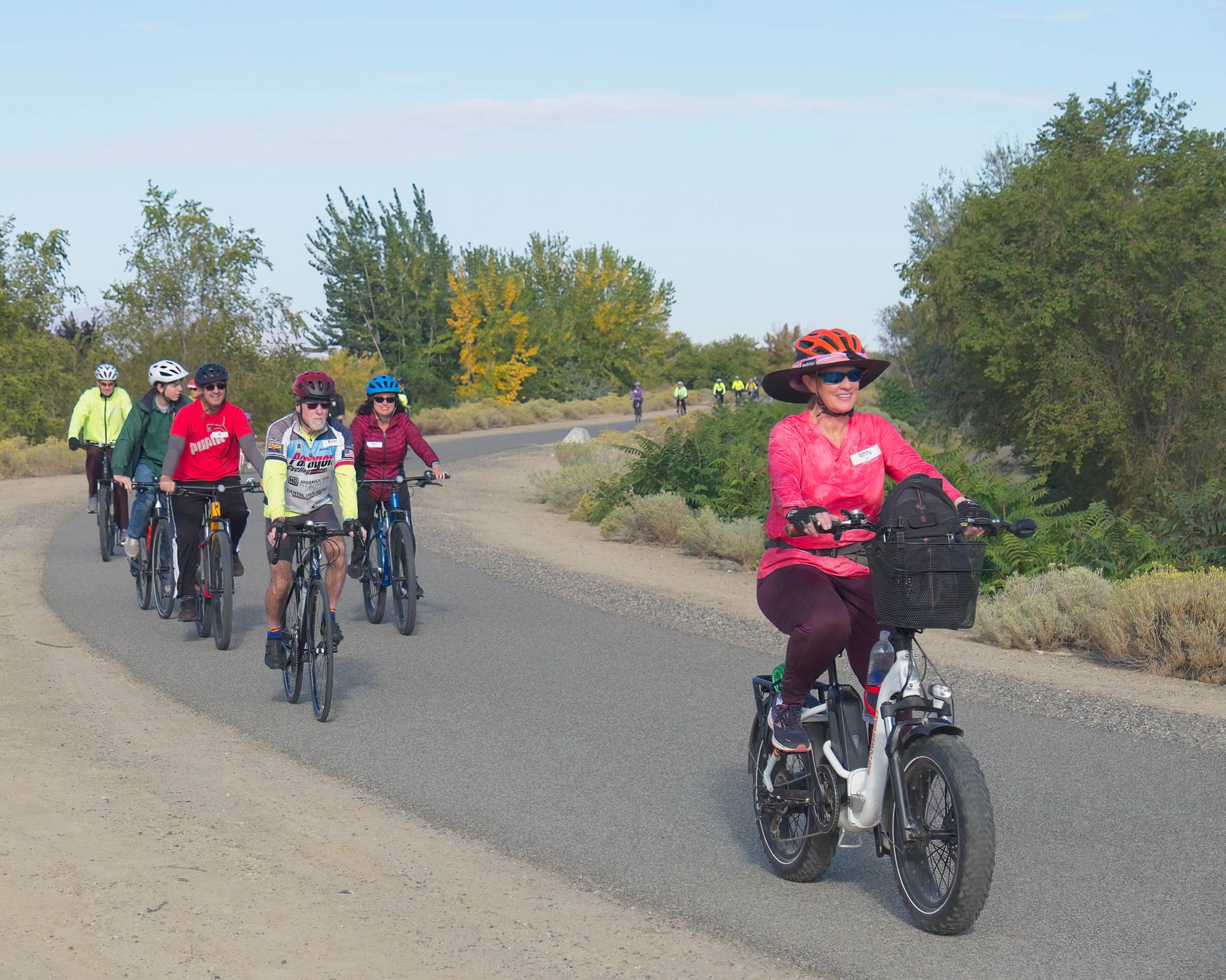 Ride with a Ranger participants enjoy a sunny day 5 cyclists on a pave path ride towards the camera on a bright sunny day. Trees with green and yellow leaves line the path and with a group of 10 riders are seen in the background.