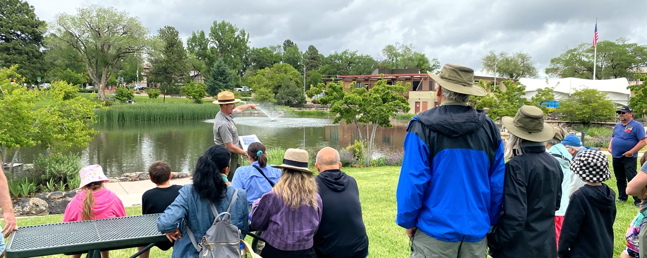 Park ranger stands in front of visitors and gestures towards a pond and building in the background.