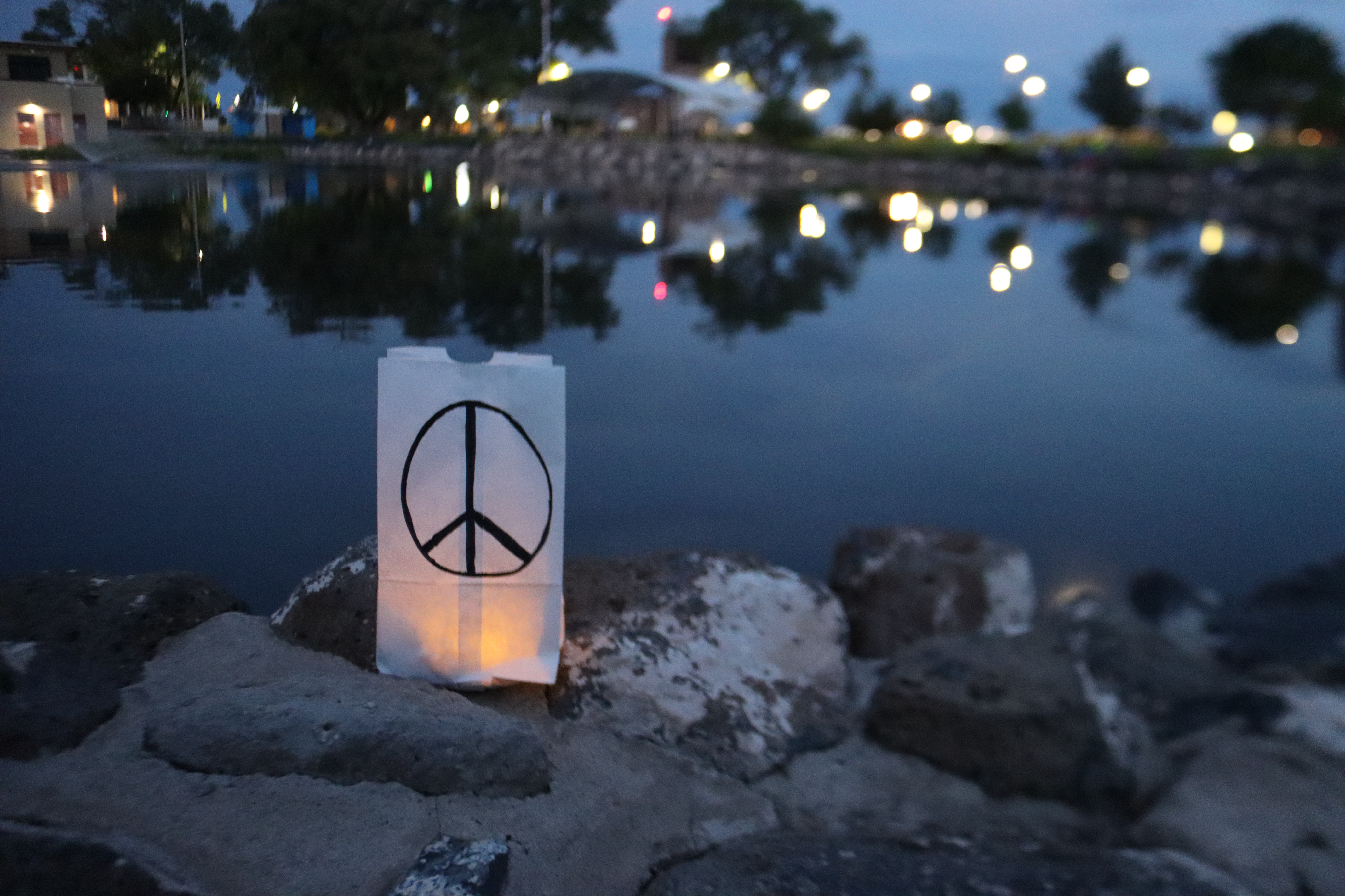 A white bag with a black peace sign drawn on it is illuminated at dusk with a lake in the background and reflecting lights.