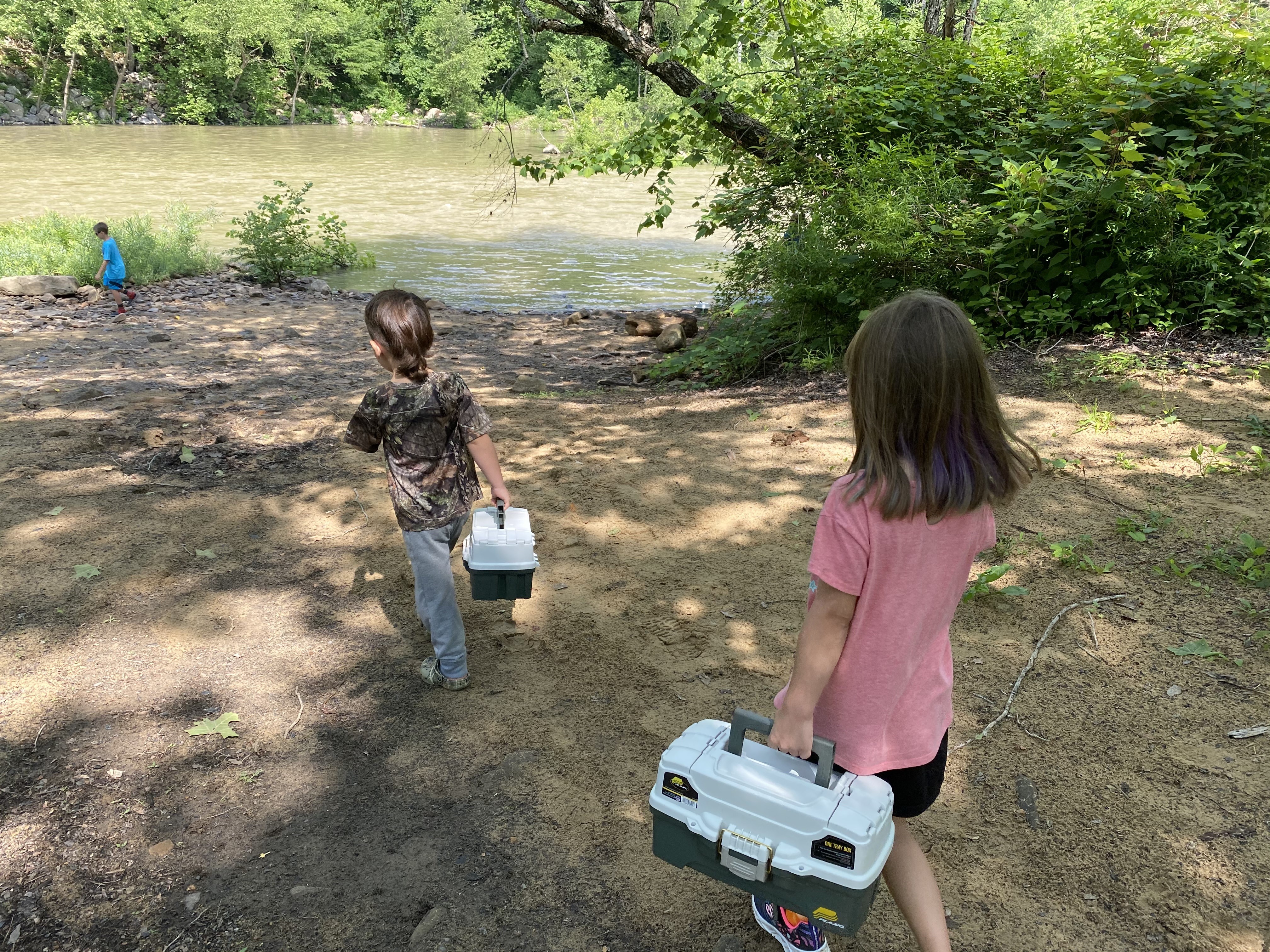 two children carrying tackle boxes walk toward a river