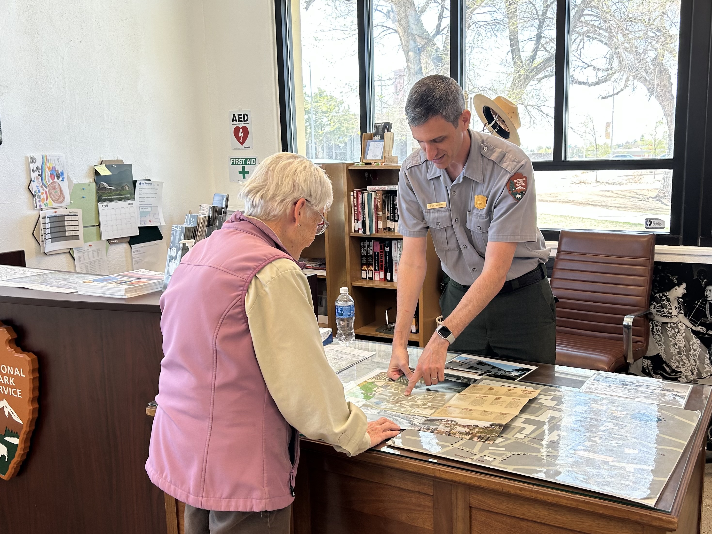 A man dressed in a park ranger uniform stands behind a desk pointing something out to a visitor.