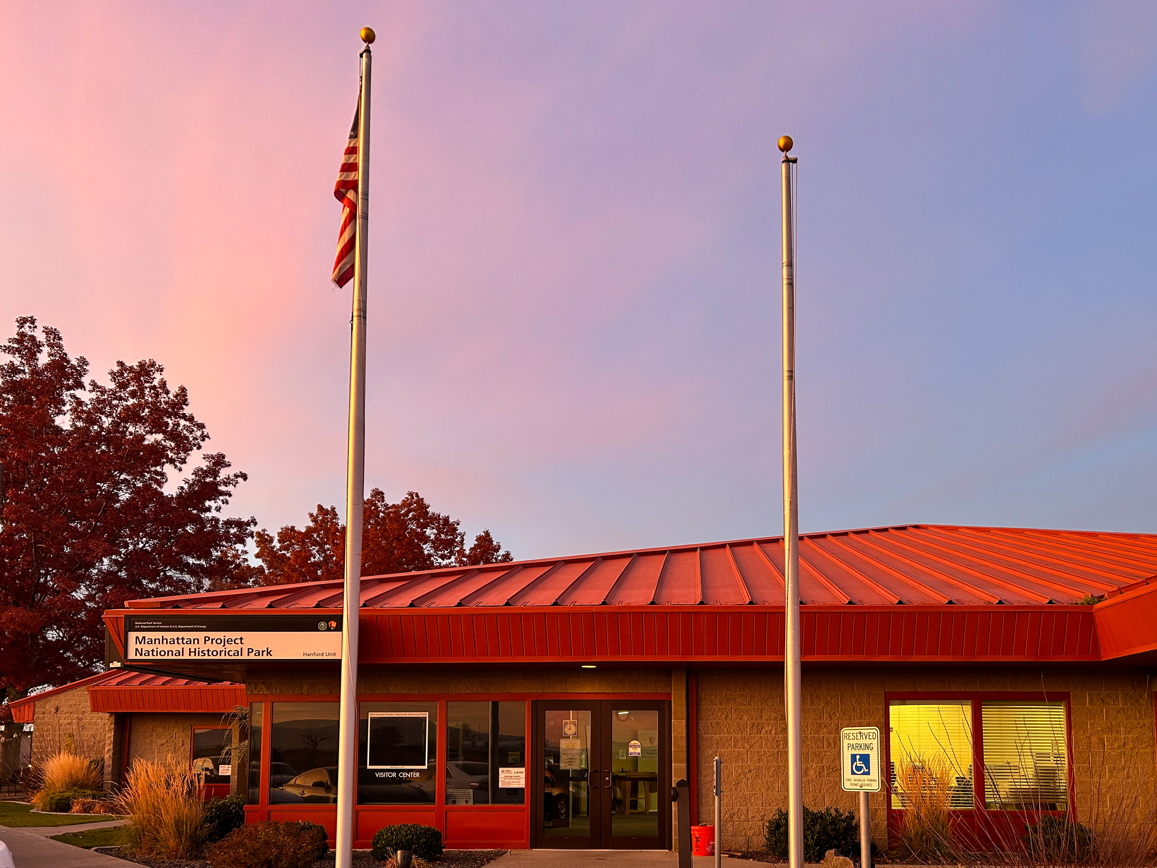 A tan brick building with a red metal roof and two flag poles in the front at sunset.