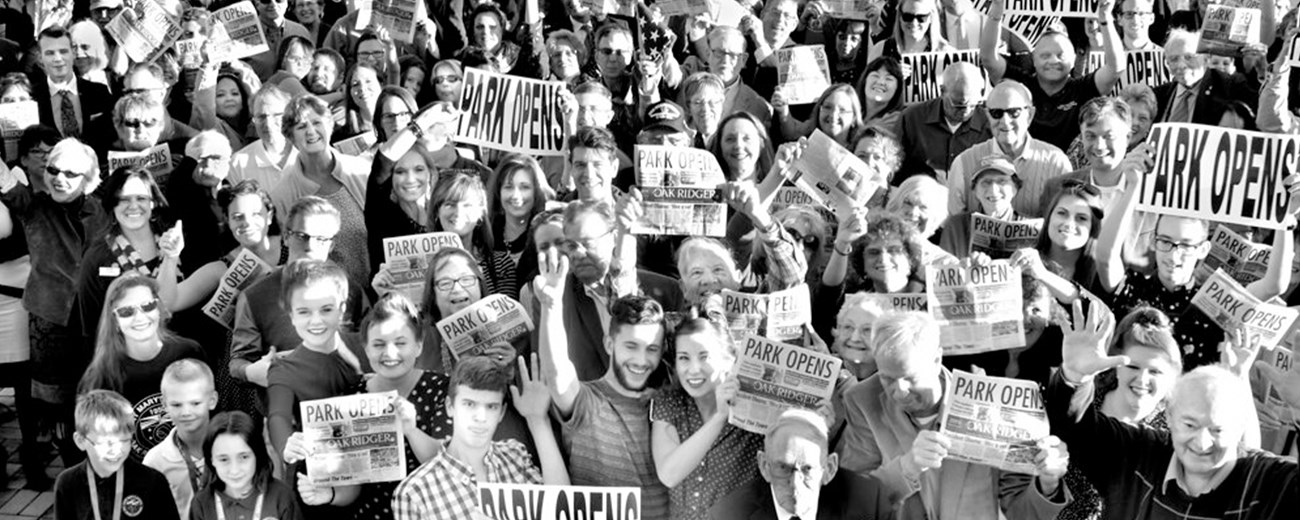 Several dozen people smile for the camera holding newspapers that read “PARK OPENS.”