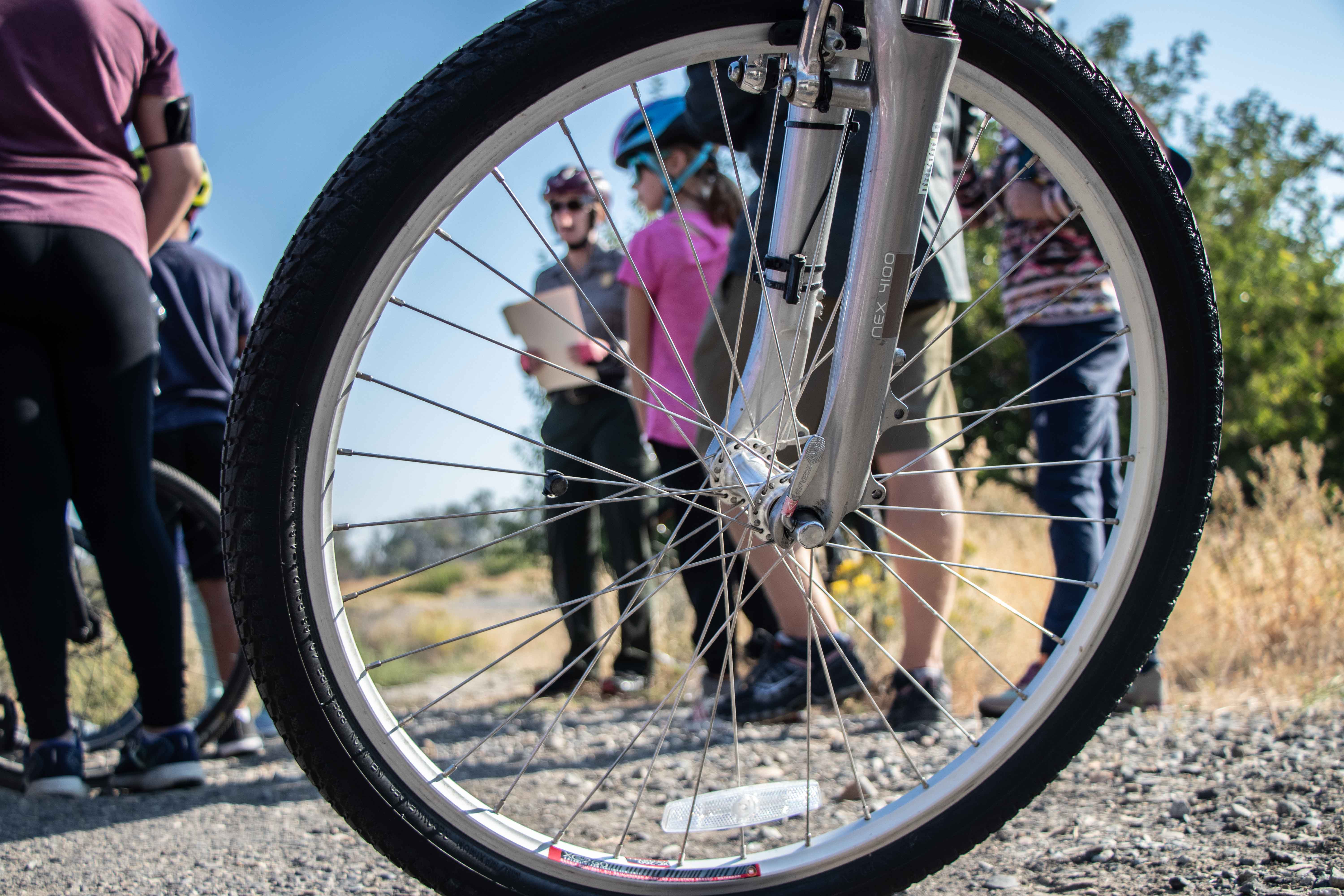The image is taken from a low angle looking through the spokes a of bike wheel with eight people gathered around listing to a speaker while standing in a field on a sunny day.