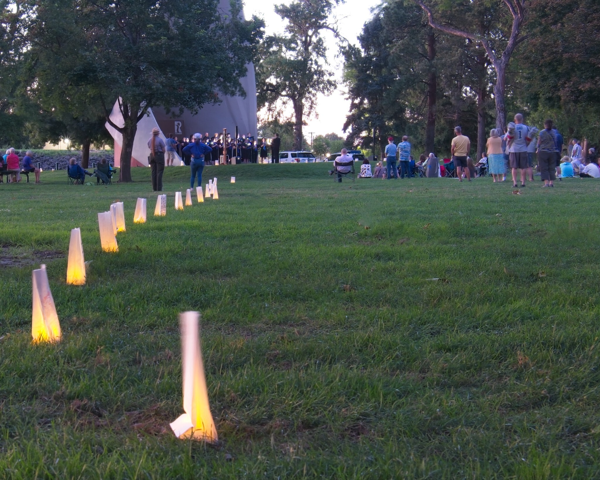 White bags in a line on green grass lawn illuminated with candles with people in the background watching a band that is performing.