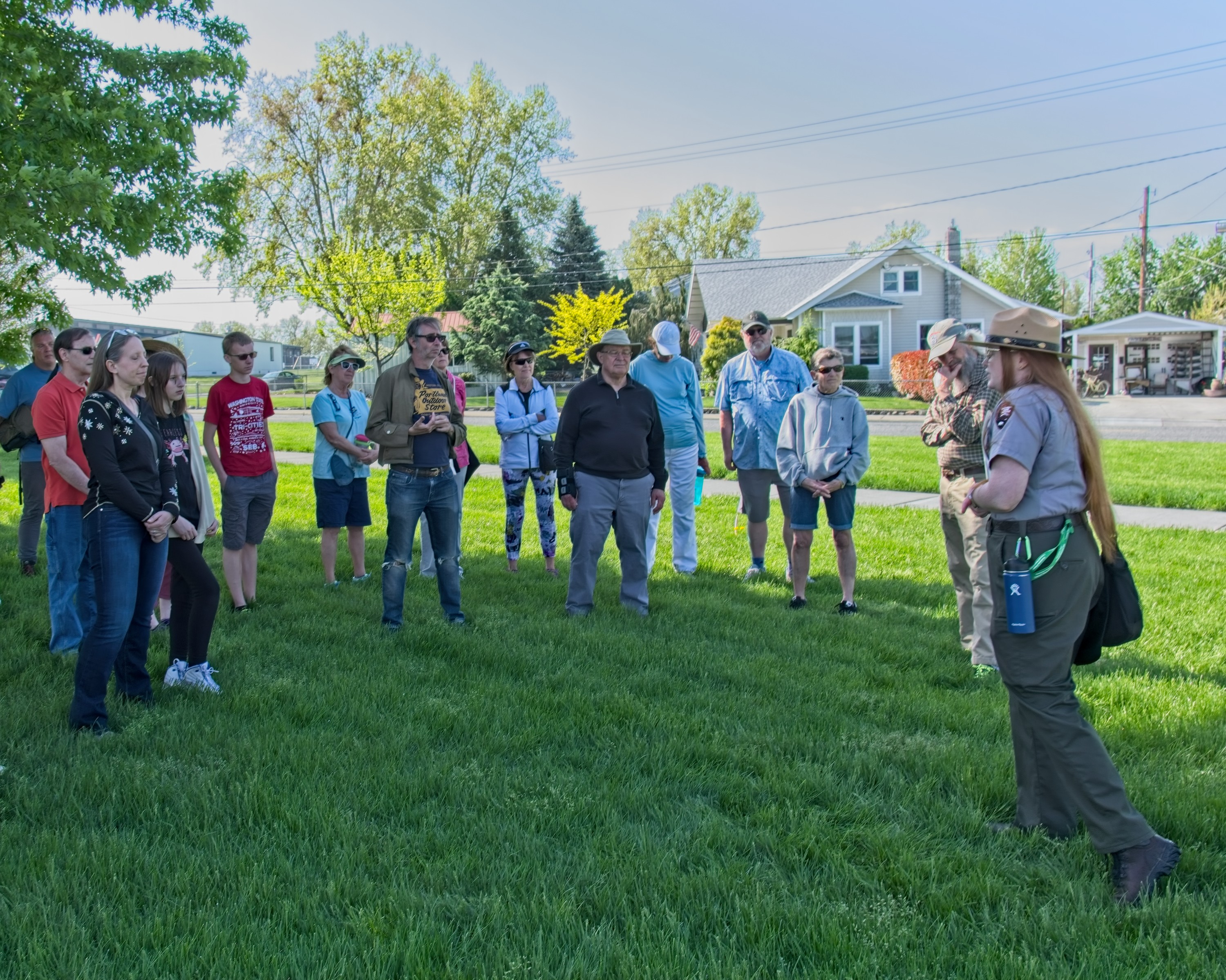 Participants learn about the history of Richland from a ranger. a group of 15 adults stand in a semi-circle on a grassy area facing a woman wearing a campaign hat, gray shirt, and green pants who speaks to the group.