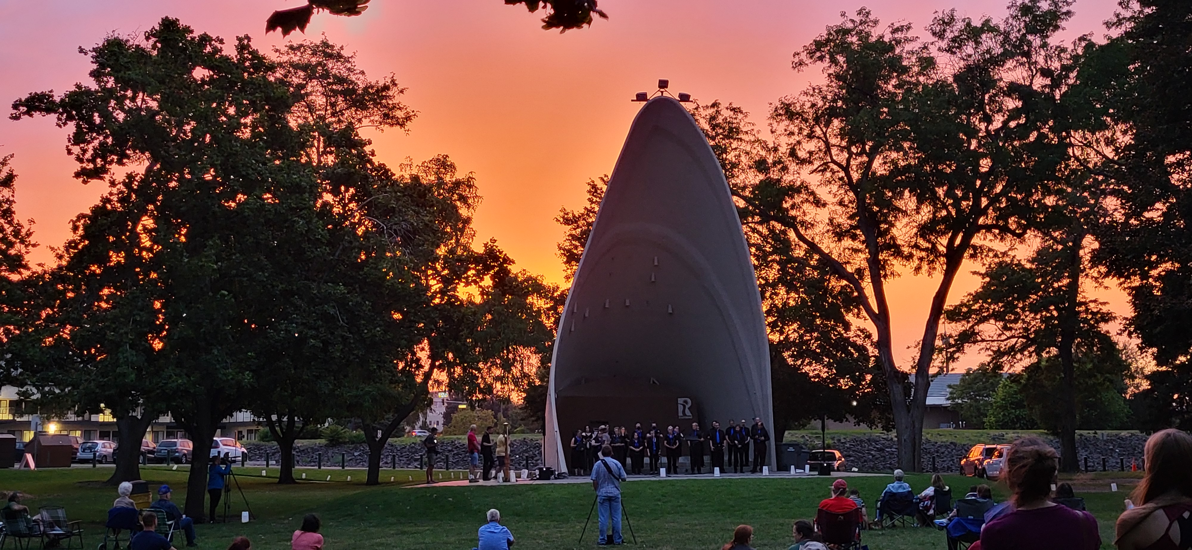 A beautiful sunset brightens the sky during the 2022 Lights for Peace program in Richland Pink clouds brighten the sky behind a very tall and narrow 3-sided shelter with people gathered in front.