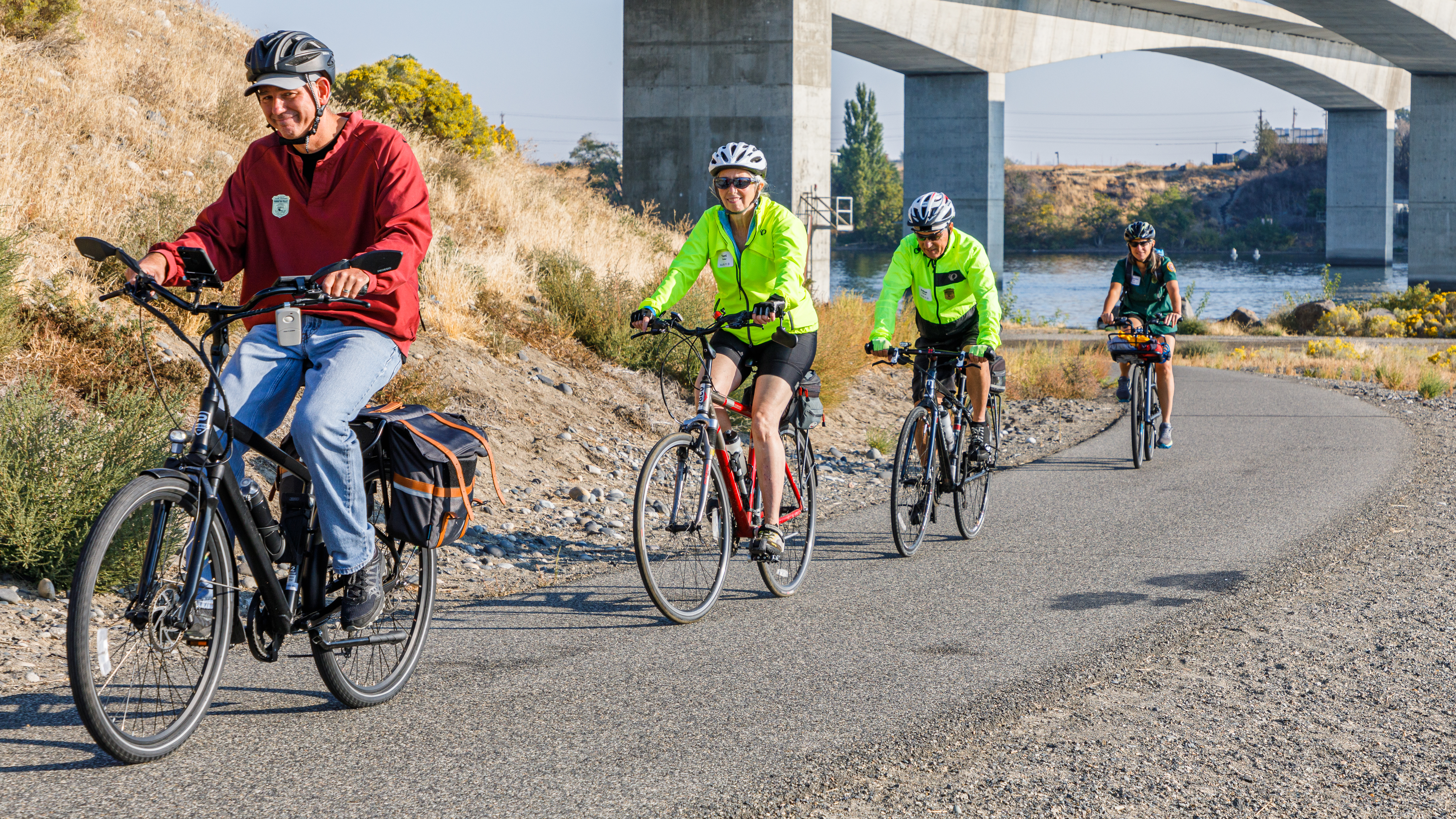 Ride with a Ranger 1 man and 3 women ride bikes in a line along a flat paved path on a sunny day
