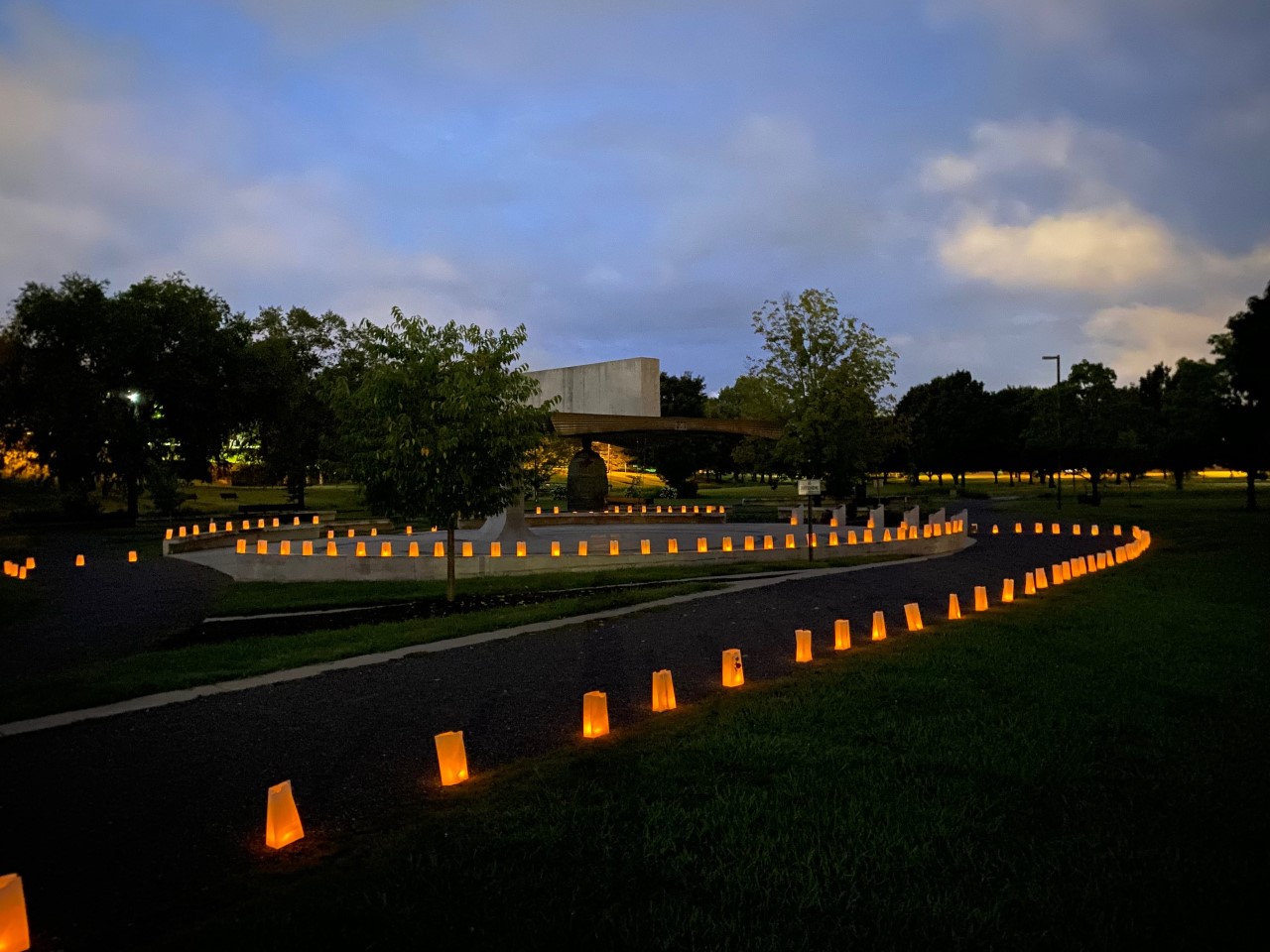 Luminarias line a path around a large bronze bell in a city park.