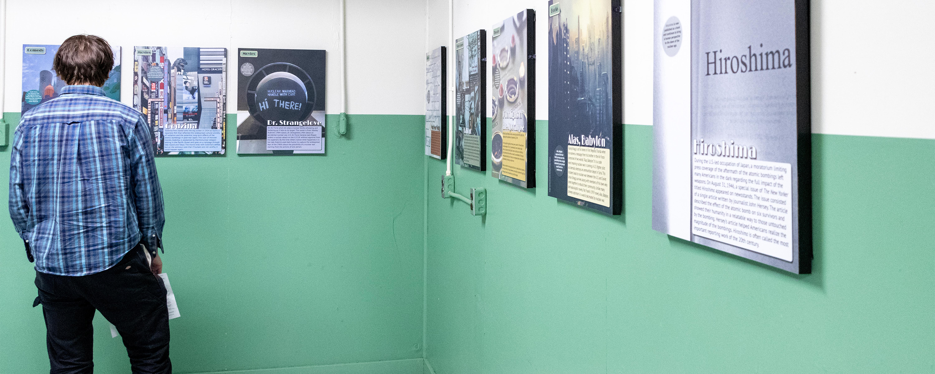 A man in a blue plaid shirt reads exhibit panels on a white and green wall.