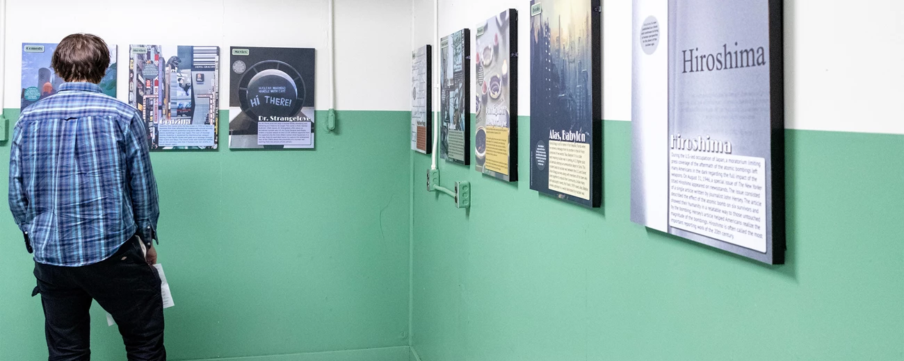 Atomic Culture room A man in a blue plaid shirt reads exhibit panels on a white and green wall.