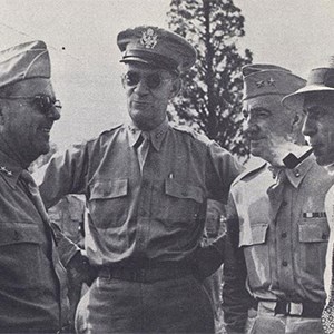  A black and white photo of four men, three in military uniform and one in a white shirt and tie, stand in a close semicircle. The man in the white shirt has a lit pipe in his mouth.