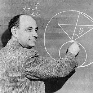 A black and white photo of a balding, middle-aged man in a gray suit drawing formulas on a chalkboard with his right hand as he looks to the right of the frame.