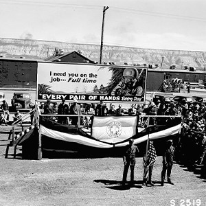 A black and white photo of several dozen people gathered at a rally. Some are standing on scaffolding while others stand along the sidewalks. Two banners are visible behind a three-man color guard in the middle of the street.