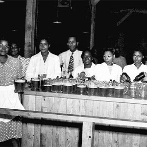 A black and white photo of several African American men and women standing at a counter. Several glass pitchers are stacked on the counter.