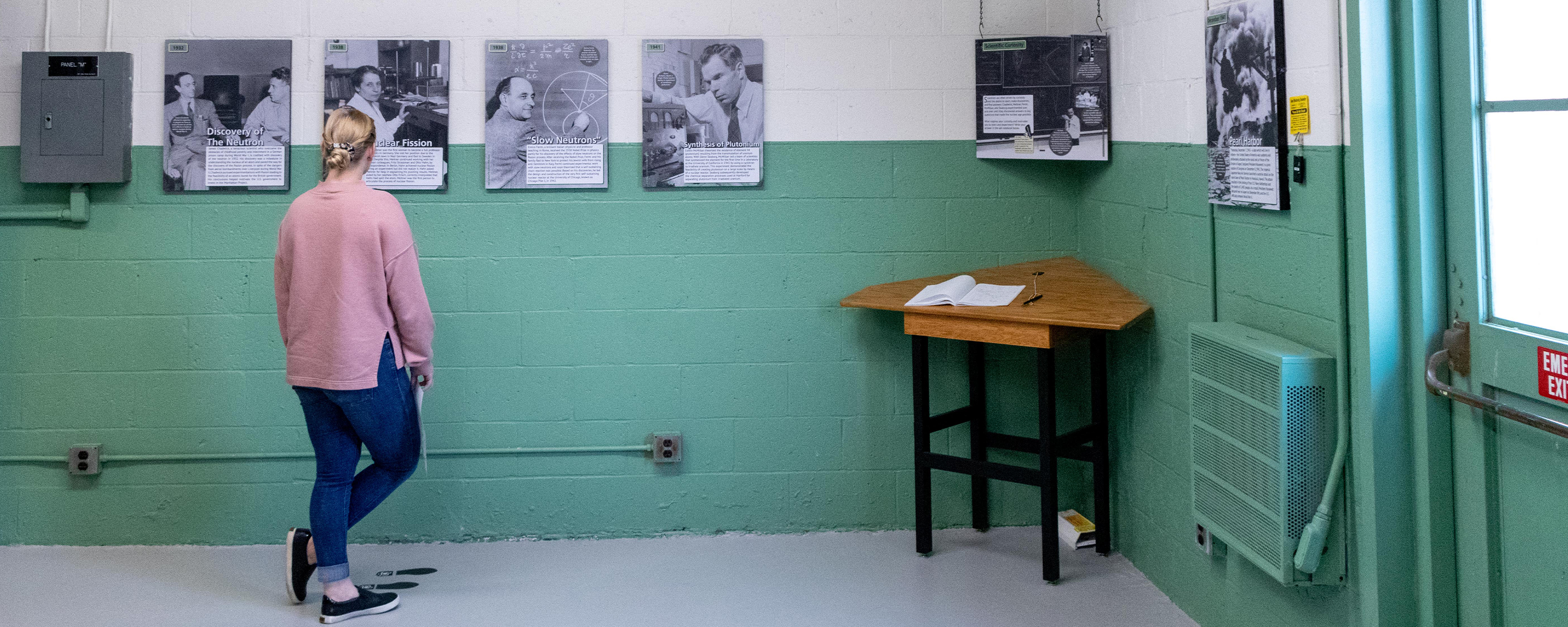 A woman in a pink shirt and blue jeans reads exhibit panels on a green and white wall.