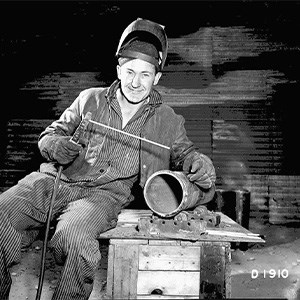 A male worker in welding gear leans in a chair and smiles for the camera.