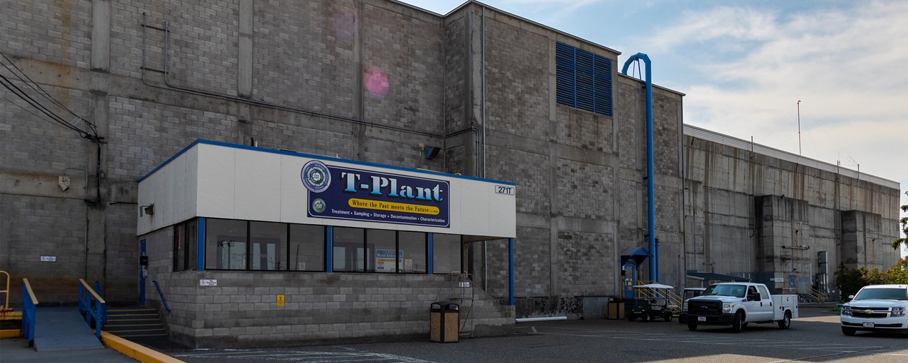 A long rectangular 2-story concrete building fills the frame and contrasts with the bright sun and blue sky. Several cars parked in front of the building.