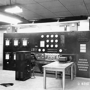 A black and white photo of the reactor control room desk, with a seating area and multiple knobs, dials, and levers all over the desk and nearby walls.