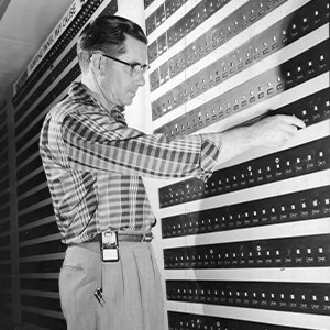 A black and white photo of a middle-aged man in a plaid shirt pushing one of hundreds of small buttons along a wall.