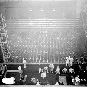 A black and white elevated photo of a tall reactor face. Approximately one dozen workers are seen standing on the floor at the base of the face.