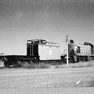 A black and white photo of two train cars next to each other in the desert. A man in white stands on the train car at right. A railroad crossing sign is in the foreground between the two cars.