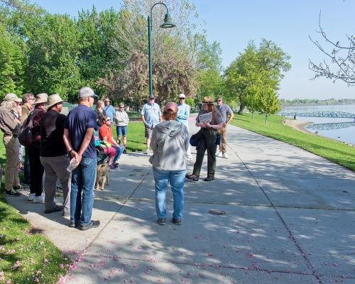 A ranger in uniform speaks to a group of people on a sidewalk near a river.