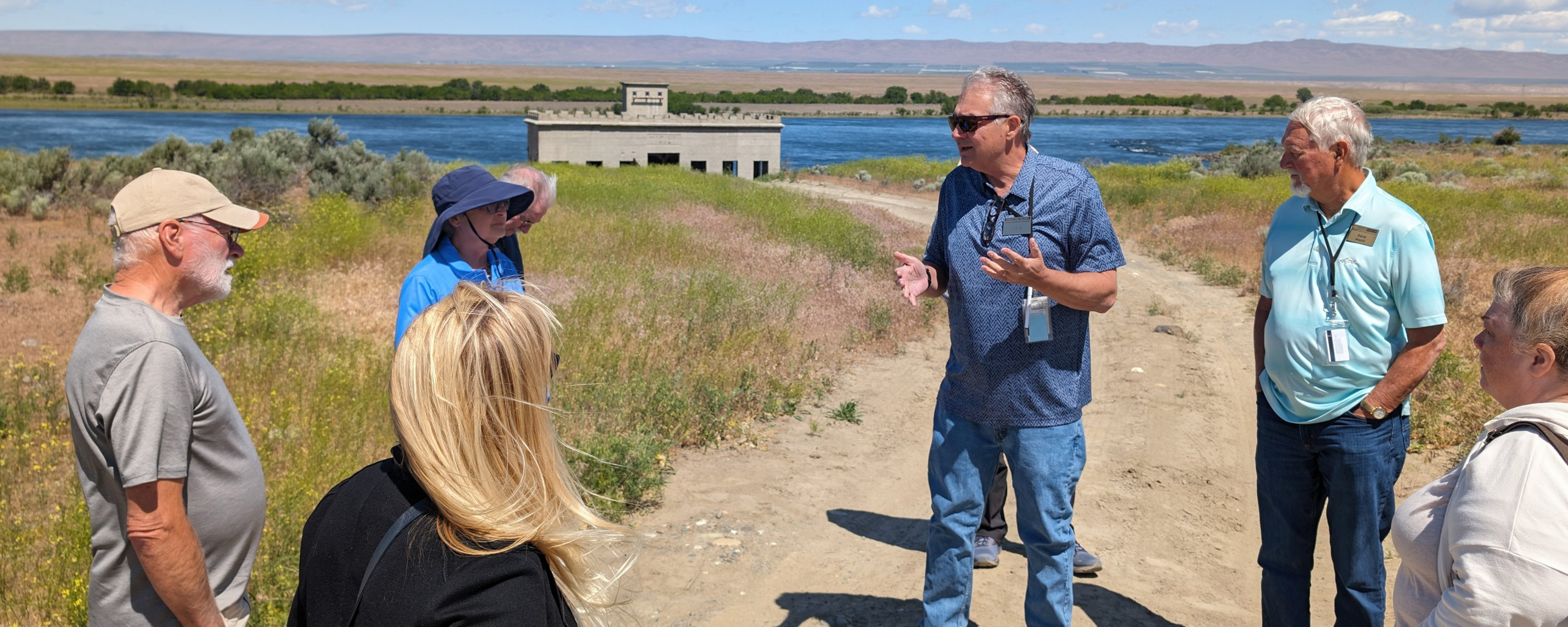 A group of people listen to a man who's speaking with a building in the background.