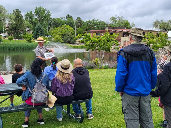 A ranger in uniform stands in front of a group of people pointing at resources he's displaying in his hands. A body of water is seen in the background.