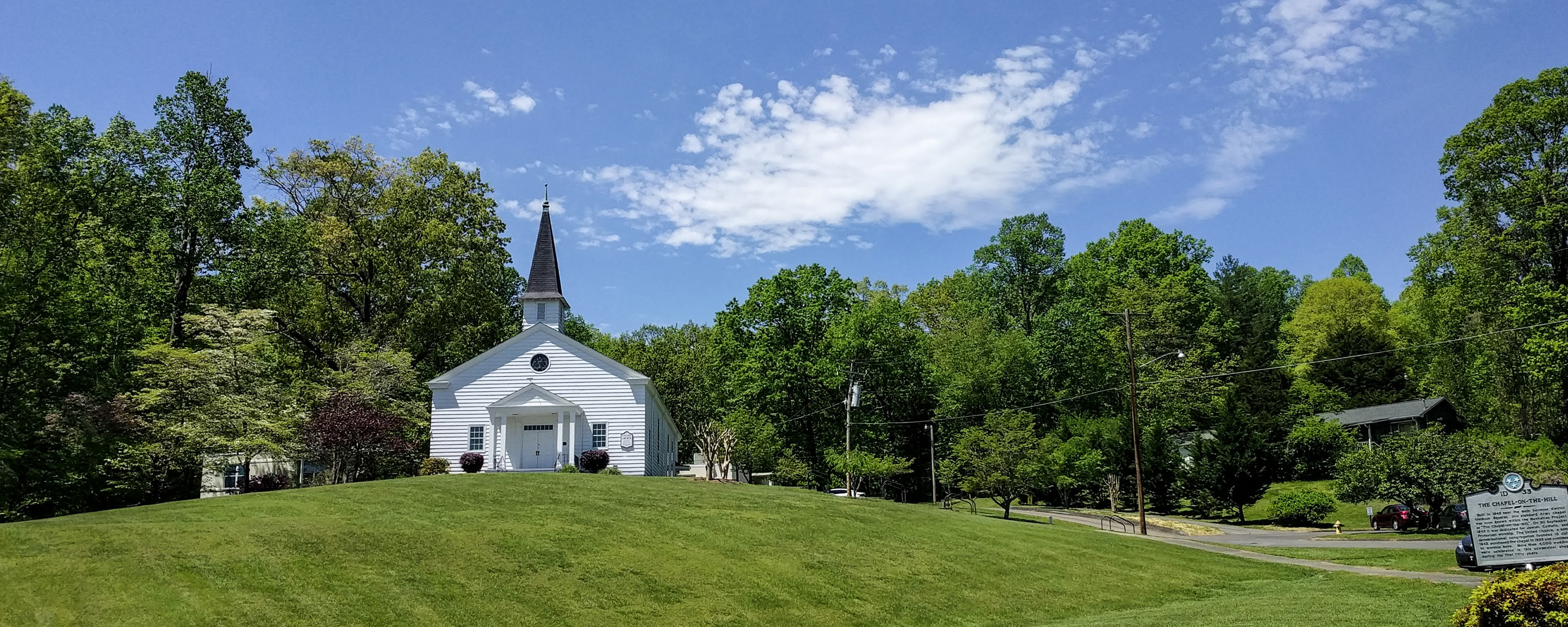 A green grassy hill with a white chapel at the top surrounded by trees. A historic sign can be seen in the foreground.