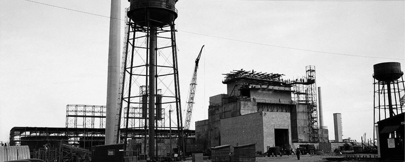 A large building under construction with a water tower and smokestack at left.