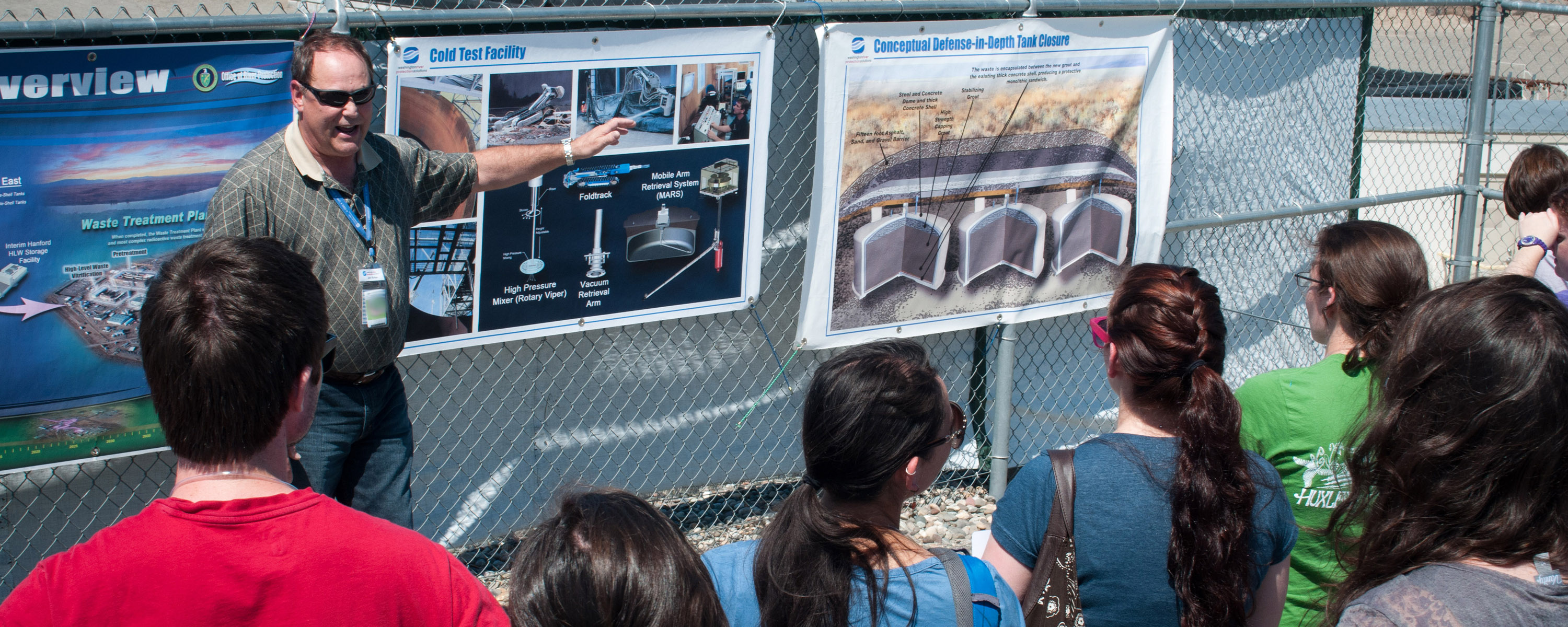 Several students gather outside as an instructor points at a sign on a fence.