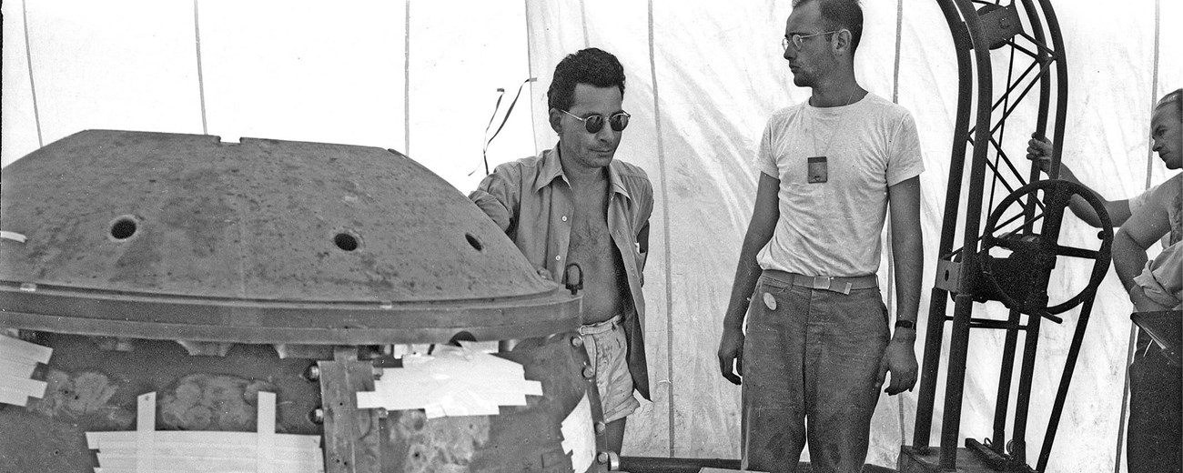 Three men surround a large metal orb inside a white tent.