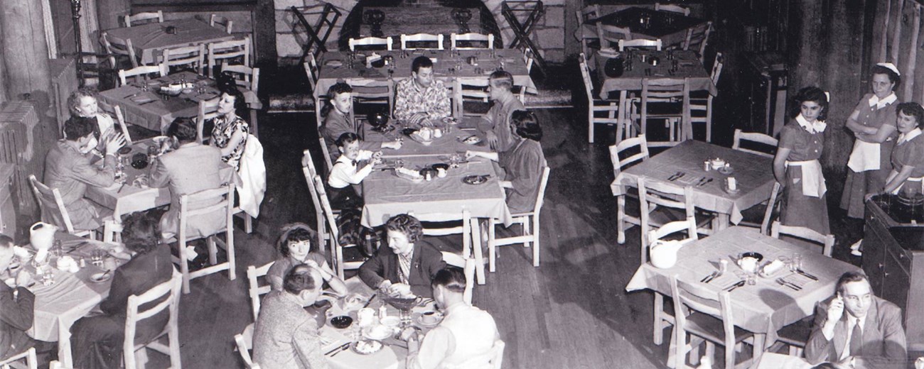 Several people gather at dining tables inside a wooden lodge with a large fireplace on one wall.
