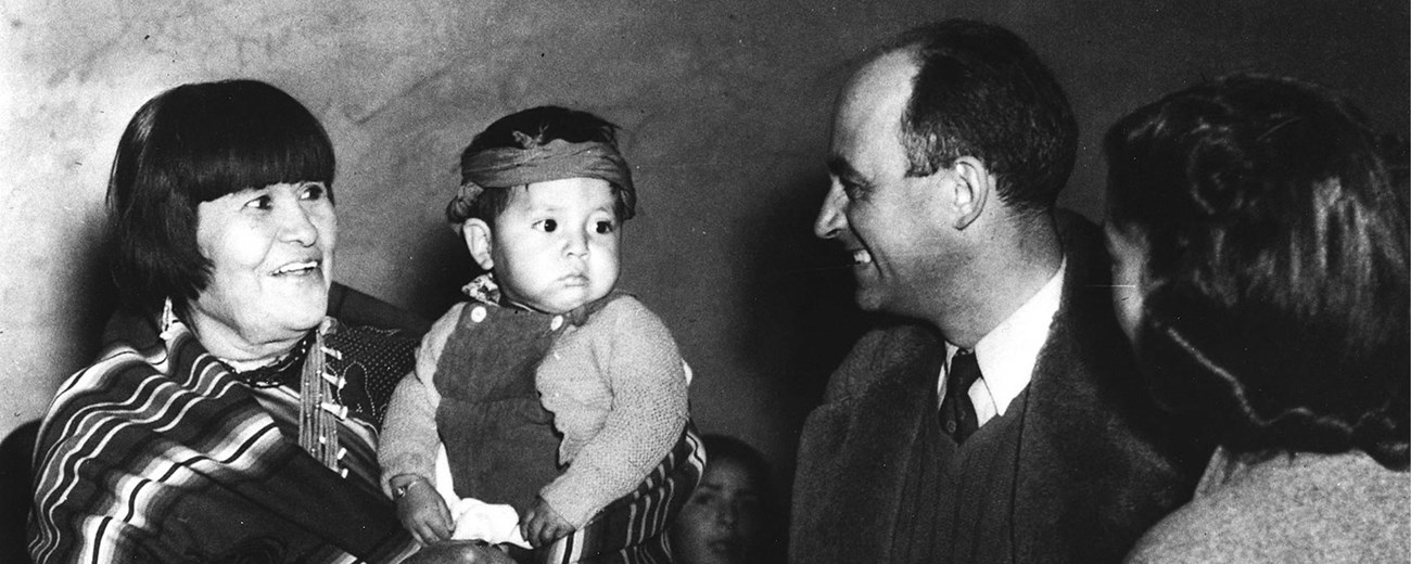 A Native American woman holds a small child as two other people look on.