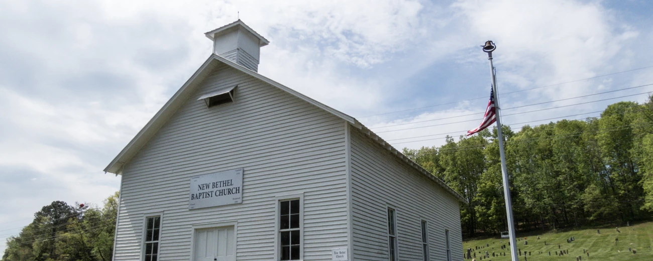 New Bethel Baptist Church The top half of a wooden white chapel with green grass and grave stones in the background.