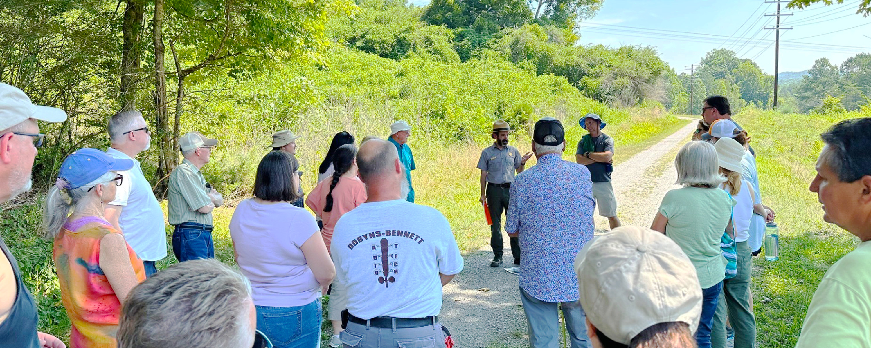 A group of people face away from the camera looking at a park ranger who is speaking. There is grass and trees in the background.