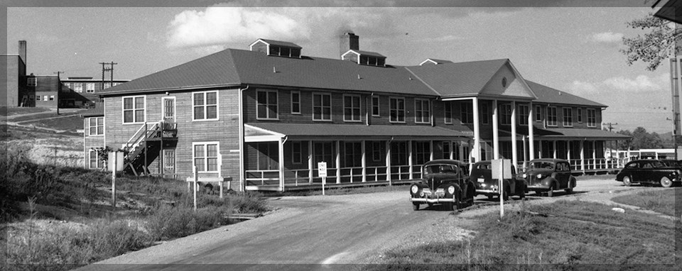 Black and white photo of a building with many windows and at least two stories. There are cars parked on a road in front of it.