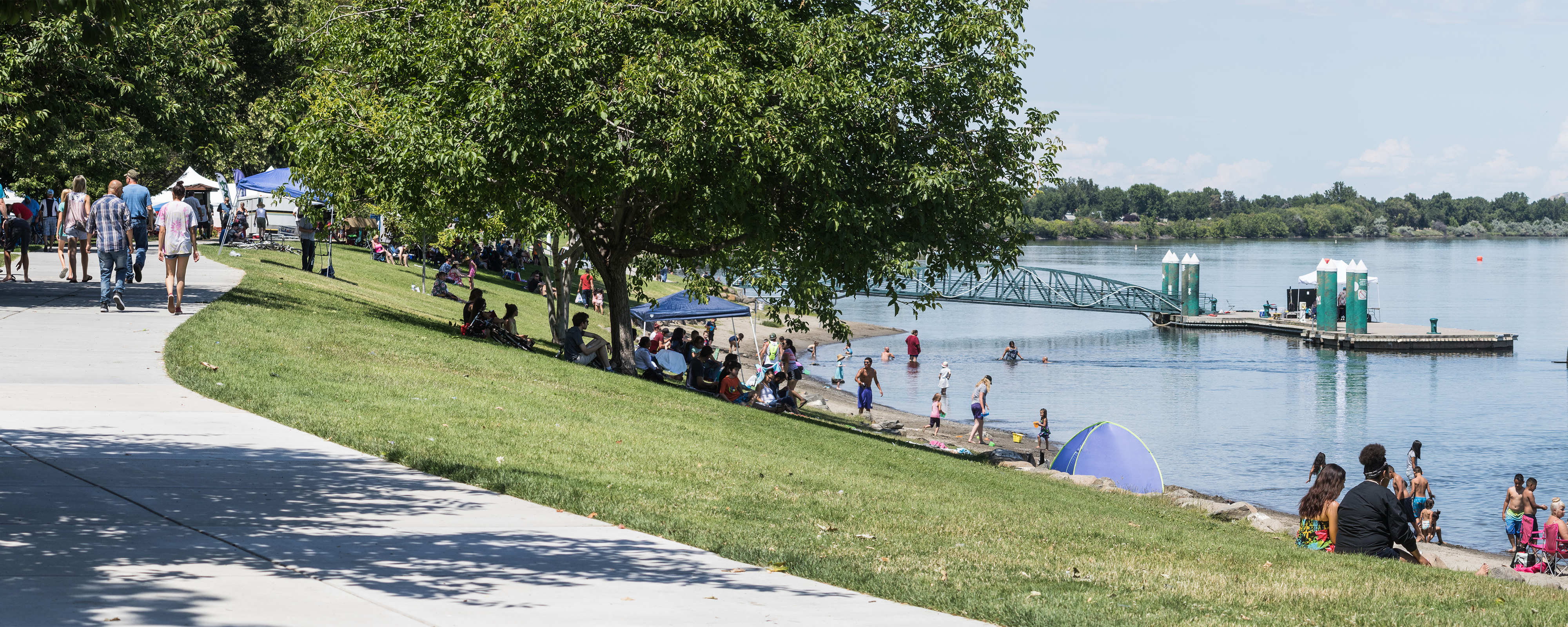 Paved path, grassy field sloping to blue river, lots of people enjoying the sunlight.